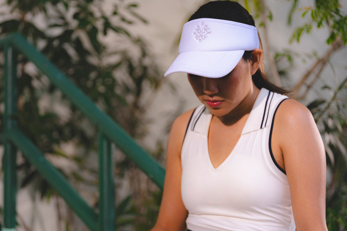 A woman wearing a white visor with a small embroidered logo and a white athletic tank top with black trim, photographed outdoors with blurred greenery and a staircase in the background. Adjustable sport visor for tennis and pickleball - Vola Face Value Visor