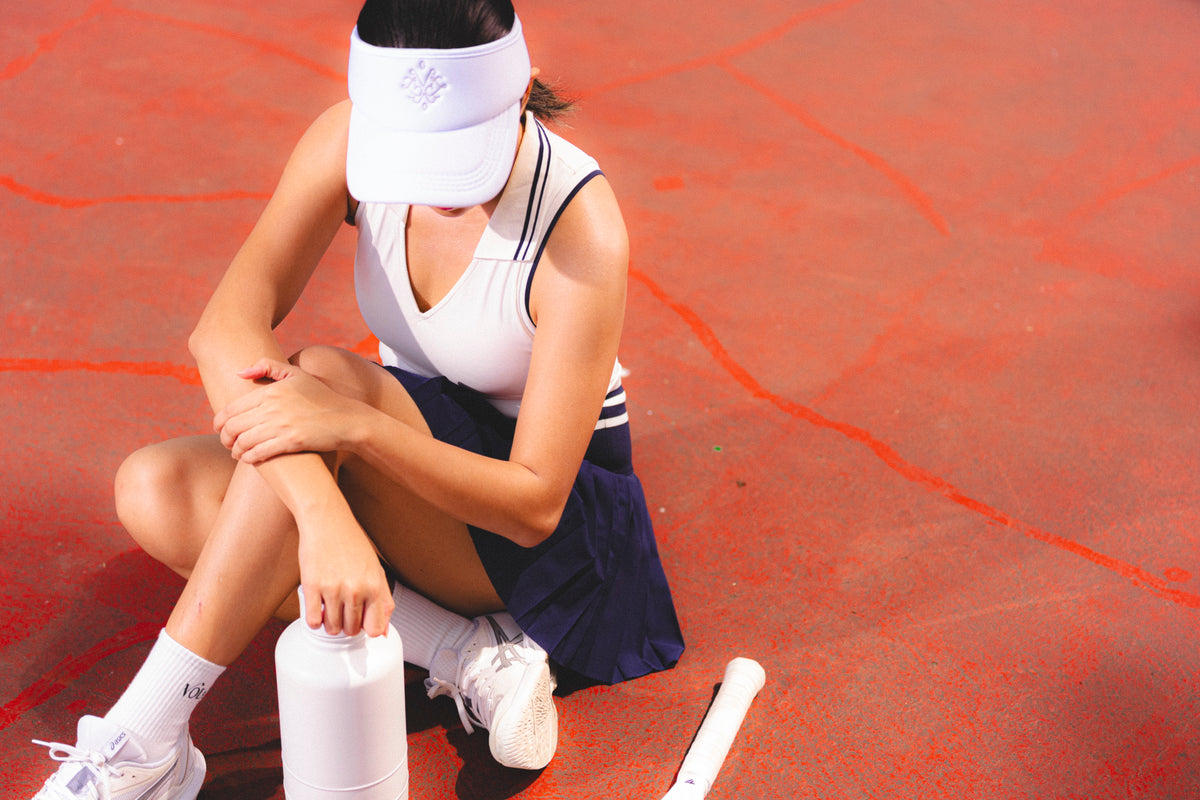 A tennis player sits on a red court surface, holding her knee and arm while wearing a white visor, white sleeveless top, navy skirt, white socks, and white sneakers. A water bottle and racket are nearby. Adjustable sport visor for tennis and pickleball - Vola Face Value Visor