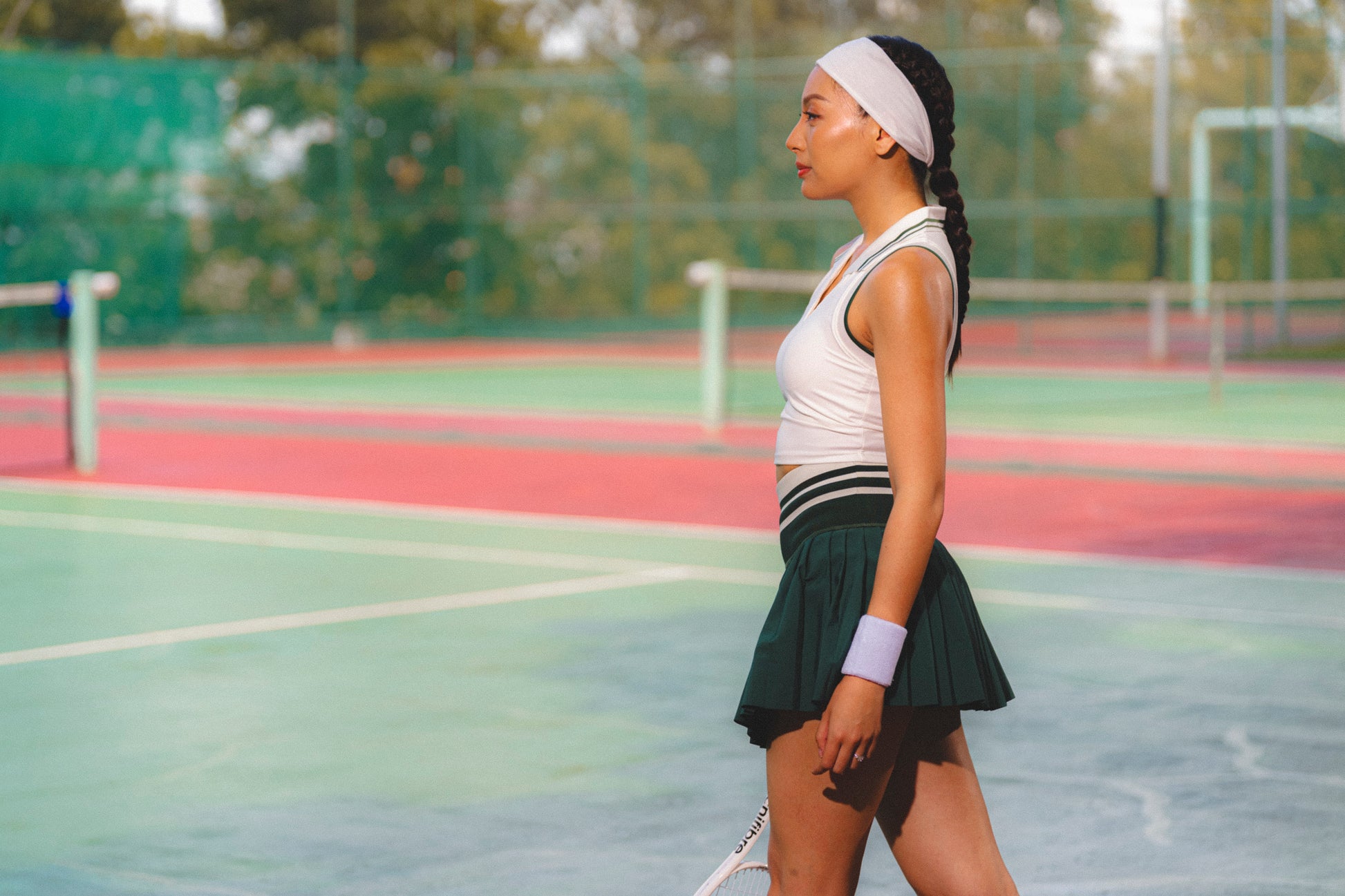 A woman in athletic wear stands in profile on a tennis court, wearing a white sleeveless top, a dark green pleated tennis skirt, a white headband, and a white wristband.
