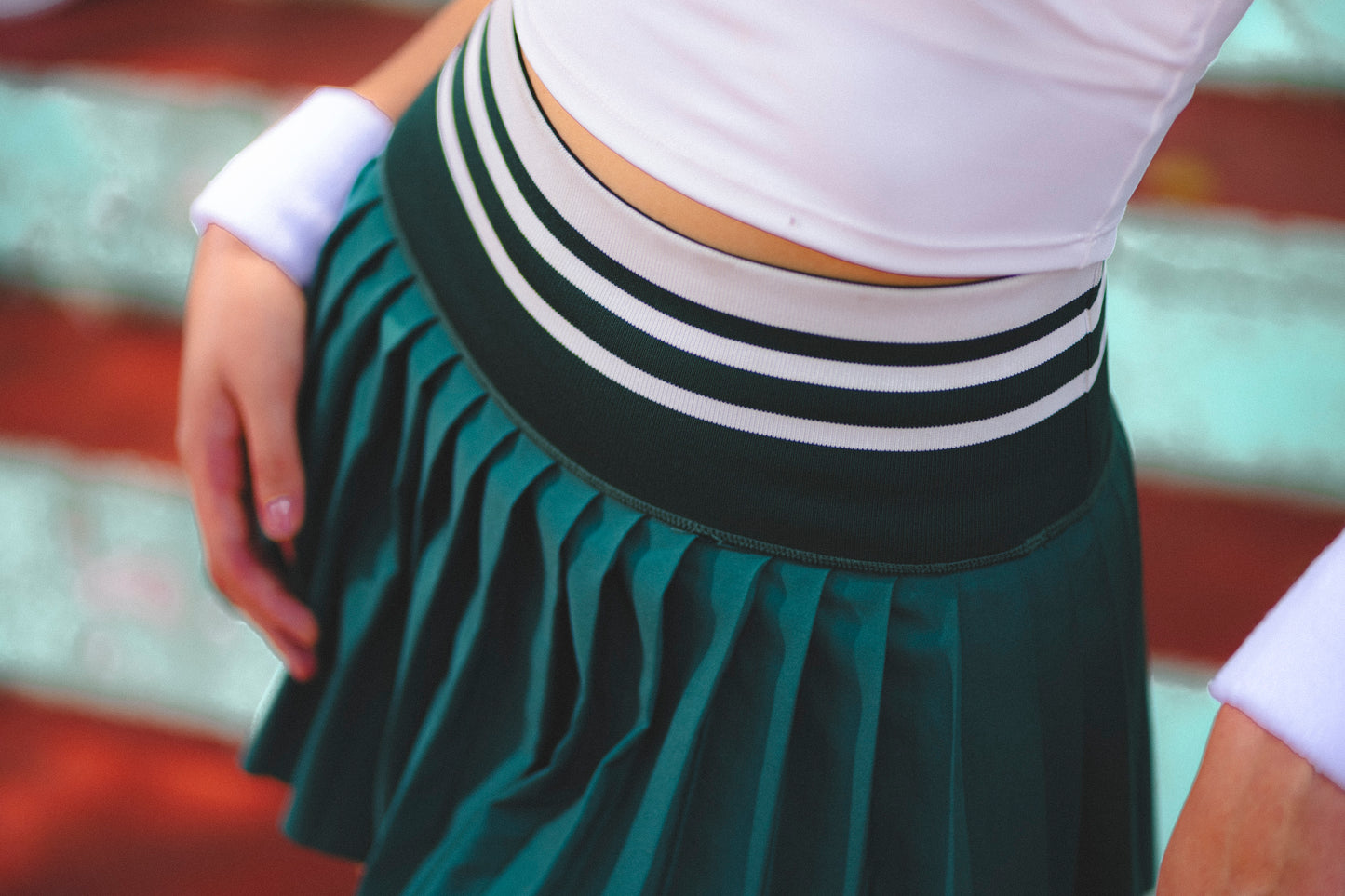 A close-up of a pleated skirt in dark green with a wide waistband featuring green and white stripes, worn with a white cropped top.