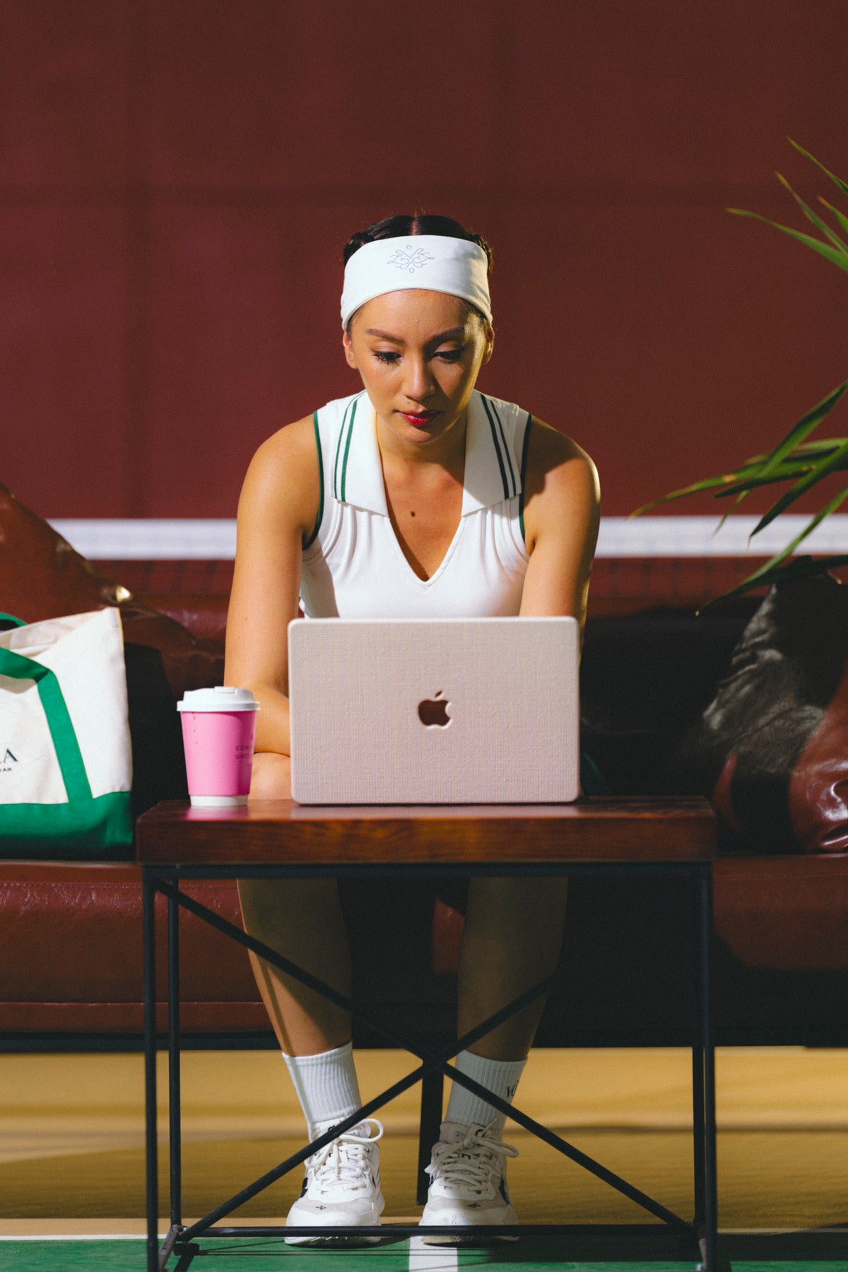 A woman in athletic wear sits at a table using a silver laptop, with a pink cup beside her on a dark indoor setting. White sleeveless tennis polo with court green collar trim - Vola Well Played Polo