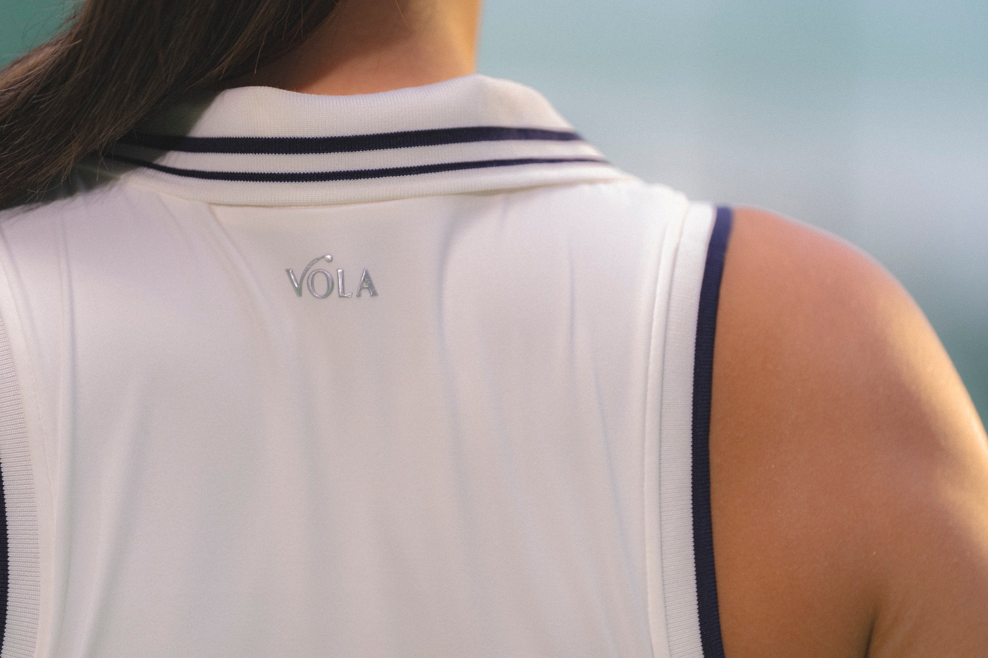 Close-up back view of a person wearing a white sleeveless athletic top with navy trim and a small gray VOLA logo centered below the collar. White sleeveless tennis polo with navy collar trim - Vola Well Played Polo.